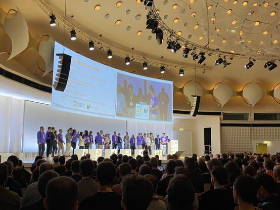 Opening Session in a large room with a big slide at top of group of pople running the conference this year.