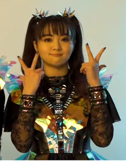 Moametal holding up both hands flashing V signs with an adorable smirk on her face.