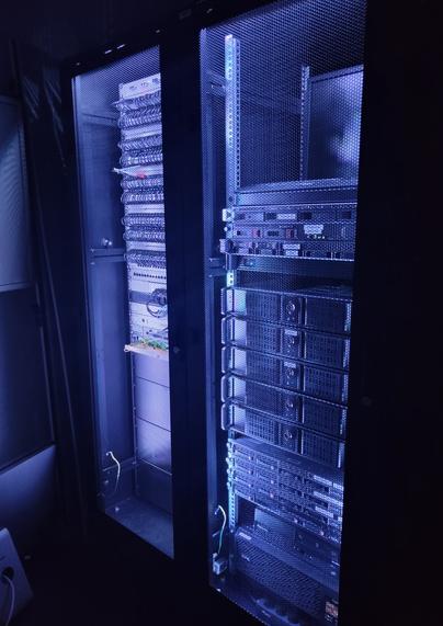 Photo of two server and networking racks in a dark room. 
The racks are lit up in a blueish purple light from the inside. 
The mesh rack doors are closed.