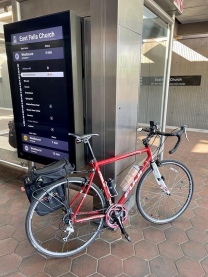 My red acoustic bike leans against a transit shelter on a train platform that has a digital display of next stops and arrivals information
