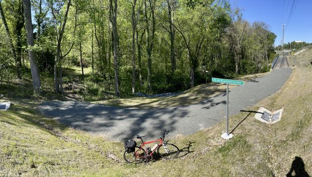 Panorama of a paved asphalt trail that ends at a gravel trail into a dirt trail leading into a forest