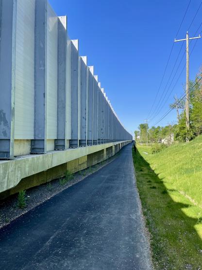 An asphalt paved trail runs in the shade of a freeway sound wall