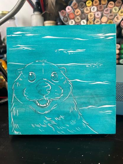 Sea otter with water ripples in the background, on a teal-colored block, with art supplies in the background.