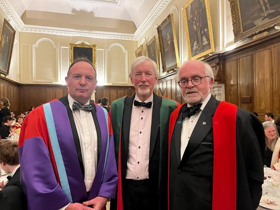 Picture of three male professors in their academic robes in a historic room with half wood panelling on the walls and a number of large framed paintings above