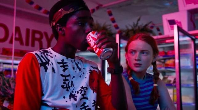 A young boy drinking from a New Coke can as a girl with pigtails looks on. They're lit by neon lights.