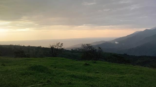 A view of the mountains from our front yard. The grass in the pastures is strikingly green, and the sky tones of orange and yellow. There's a thick layer of clouds high in the sky and the mountains are visible in the distance.