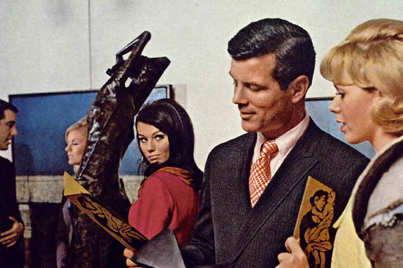 1967 A Celebrity Event from the 1960s

The image is a photograph featuring several individuals in an indoor setting.
It appears to be a group photo, likely taken at a formal event or function given the attire and the presence of name tags on some individuals.
In the center, there is a man holding a microphone towards the camera, suggesting he may be addressing an audience or announcing something.
To his left, there are two people standing; one is partially visible with a name tag and the other appears to be looking at the camera.
To his right, another individual is facing away from the camera, also with a name tag.
In the foreground, slightly out of focus, there is a woman holding what seems to be a magazine or a booklet open, possibly showing an item of interest to the group.
The environment suggests an event such as a convention, expo, or awards ceremony, indicated by the formal attire and the professional setting.
There are no visible texts within the image that provide further context about the event or the individuals present.