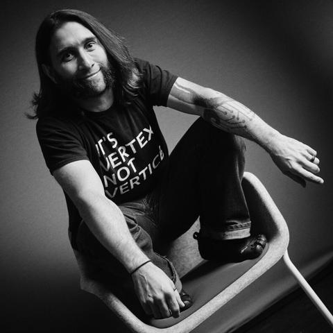 Black & white photo of a man squatting on a chair. His t-shirt reads "it's vertex, not vertice".