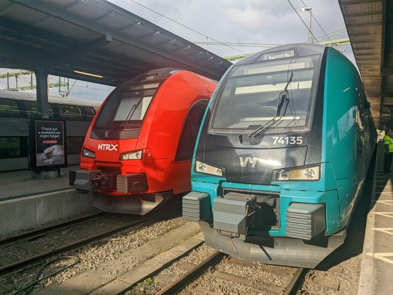 a Vy Stadler Flirt (teal livery) next to an MTRX one (red livery), with a gray double-decker in the background