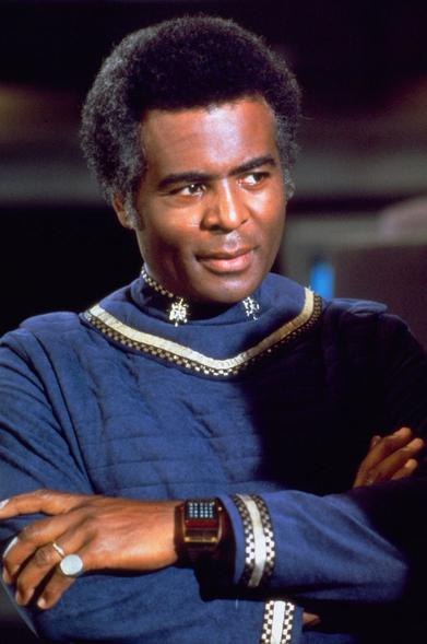 Actor Terry Carter, in character as Colonel Tigh from the original Battlestar Galactica television series. He is standing on the Galactica's bridge, in a blur officer's uniform, with arms folded displaying a high-tech watch on his wrist.