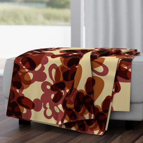 Mock-up of a haphazardly folded blanket on a white bed in front of a sliding glass door. The blanket has an abstracted pattern in reds and oranges on a pale yellow background. The design is called Forestfire Burst - Red.