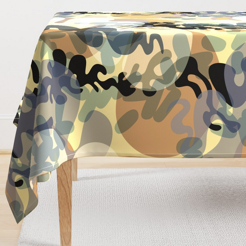 Mock-up of a patterned table cloth on a wooden table in a white room. The fabric pattern is an abstracted depiction of curls of smoke and haze intertwined with charred black squiggly shapes on a pale yellow background. The name of the design is Fire Plumes and Smoke.