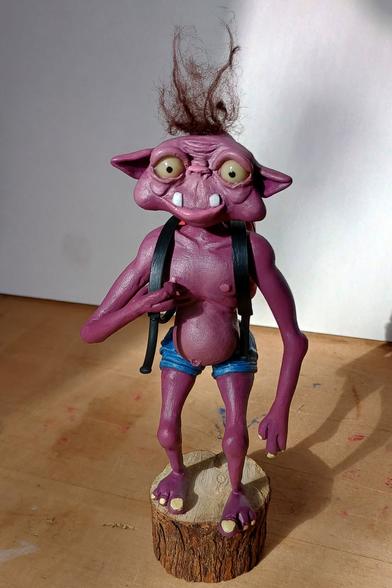 A figurine. It has purple pinkish skin, big ears, a tuft of fluffy hair, a snub nose and protruding teeth. It is wearing little jean shorts and holding one strap of its backpack.