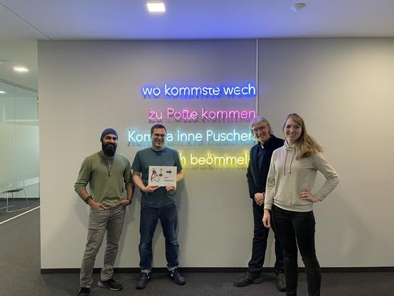 Four persons, three male, one female standing in front of a white wall. On the wall led decoration with german words is mounted.