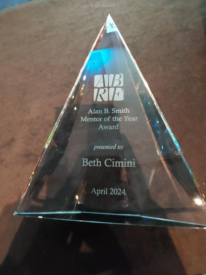 A triangular glass award saying "ABRF Alan B Smith Mentor of the Year Award presented to: Beth Cimini, April 2024"