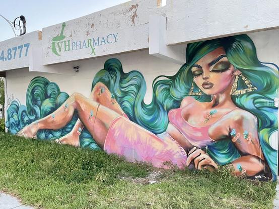Mural of a woman reclining with long flowing turquoise hair, large triangle hoop earrings, a short pink skirt, and small bandages holding turquoise flowers all over her body