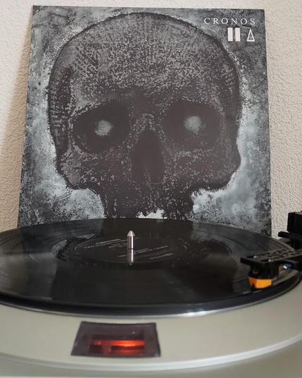 Two Fingers/Muadeep's EP "Cronos" playing on a Denon DP-30L turntable.

The cover in the background shows a stylized dark-gray and black drawing of a skull, with the artist logos and the album title in white at the top right. The record itself is the standard black vinyl.