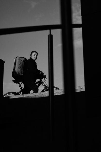 The image features a person riding a bicycle, a delivery person with large delivery backpack branded with "Glovo." The photograph is captured in a monochromatic tone, contributing to a raw and candid aesthetic. The cyclist is silhouetted against a brightly lit sky, and we observe this scene framed by a dark, vertical structure, that effectively splits the image into two halves.