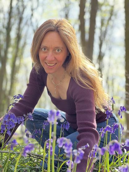 woman almost sitting (in a pretty way) near the ground in the woods, with bluebell flowers all around her and tall trunked trees in the background. She is smiling and her medium long red-brown hair is looking pleasant. She's wearing a burgundy long sleeved top and jeans.