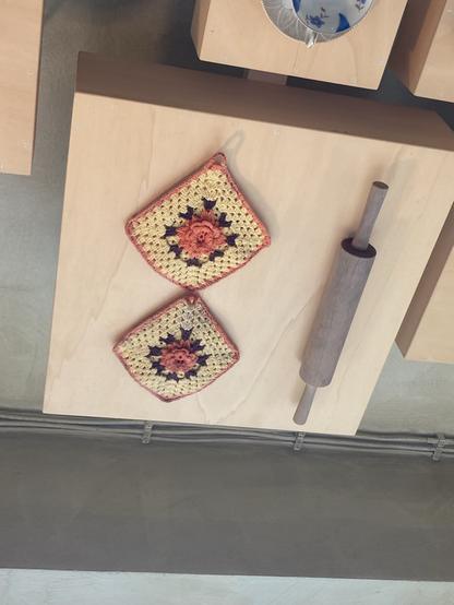 Deckenplatte aus Holz, daran befestigt 2 gehäkelte Topflappen mit mittiger Blumenrosette. Teigausroller.
