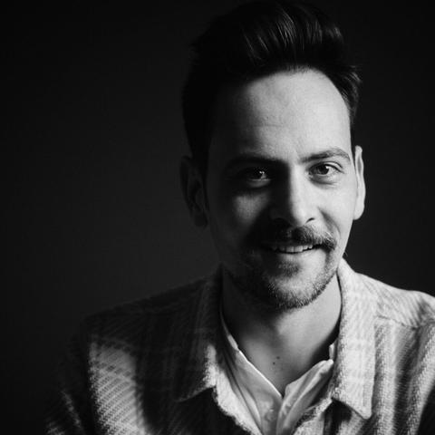 Square, black & white photo of a moustached man smiling into the camera.