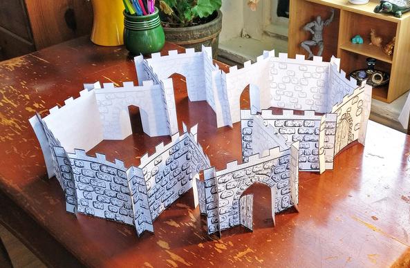 A photo of a roofless paper castle printed with black ink on white paper, sitting on a scratched reddish brown wood desk. The castle has two hexagonal rooms to either side, with a hallway/double foyer area in between that is long from front to back, but narrow between the hexagonal rooms. The nearer end of this hallway has 3 open doors, and the far end has one. There are doors on both hexagon rooms as well, some open and some closed. All the doors are pointed gothic arches.