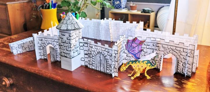 A photo of a large roofless black and white paper castle and a yellow and purple paper dragon arranged on a scratched wooden desk. The castle has one narrower section on the right behind the dragon. The chamber on the left is a large square with two large doors on the front. On the wall between the doors is a tower with a conical roof. An unused castle part sits to the left of the castle.