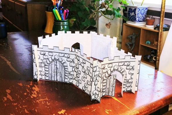 A photo of a roofless black and white paper castle arranged on a scratched wooden desk. The front of the castle is a small square chamber that opens into a larger hexagonal chamber in back. There are three doors with pointed gothic arches on various walls of the castle, two of which stand open.