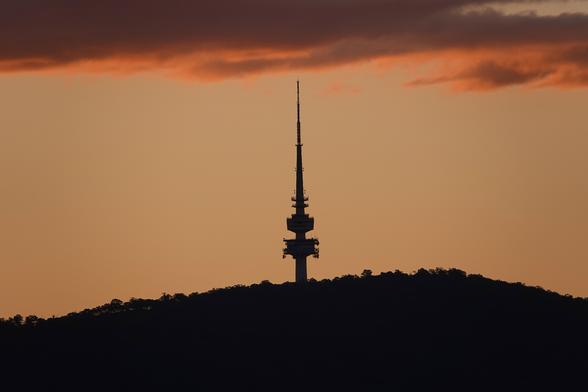 Black Mountain (Canberra) at sunset. The sky is orange with purplish clouds at the top of the image. The view is a silhouette of the top of a hill with a needle-like telecommunications tower at the top.