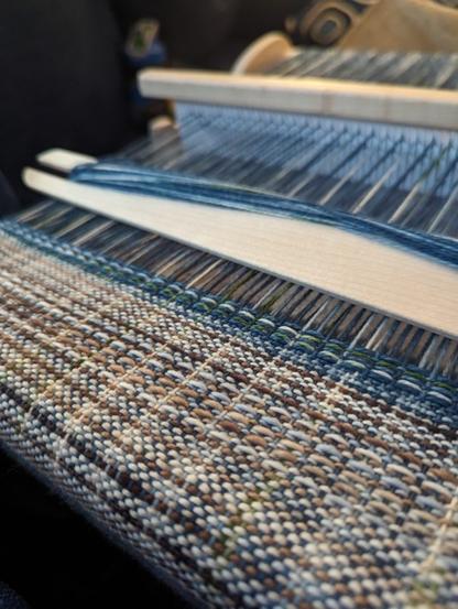 A cloth in process of being woven on a rigid heddle loom. There are a few sections with short weft floats just woven in the lighter grey-brown and then more done in a blue-green yarn.