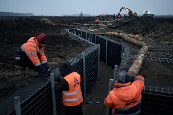 Photo of construction work in a war zone in Ukraine. 

It's an open area. No trees, bushes or buildings. A zig-zag trench is being dug with an industrial digger. The trench has metal rods for reinforcement in prep for possible concrete to follow.

All the workers are wearing high visibility vests or jackets.  Remember they are 100% out in the open in the middle of a war zone.