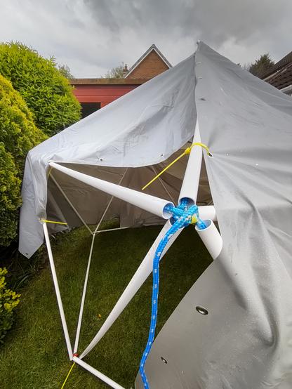 Photo of a very basic 1V geodesic dome made of white plastic tubing and blue and white braided rope. It is partially covered by two heavy duty grey groundsheet "tarps", attached with bright yellow cable ties.