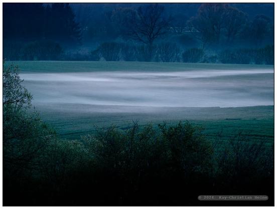 Bodennebel steigt in der blauen Stunde am Abend aus den nassen Feldern auf und überzieht das Land in der Dämmerung mit einem silbrigen Schimmer, betont durch die raffende Wirkung eines Teleobjektivs.