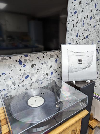 Photo of the clear vinyl "Shape Of Fluidity" playing on a turntable with the sleeve propped up to the right. The sleeve is a black and white photo of a (plastic?) flag dripping water droplets