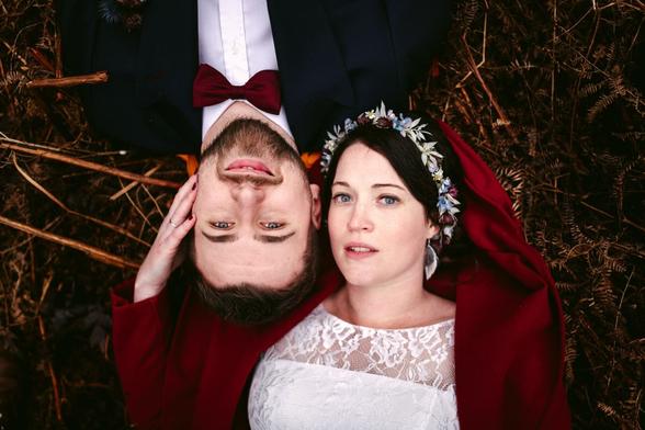 The photo is a top-down view of a bride and groom lying side by side on a bed of rust-colored pine needles. The groom is wearing a classic dark blue suit with a white shirt and a red bow tie, while the bride is adorned in a traditional white lace wedding dress with a floral headpiece woven with small flowers. She is also wrapped in a rich red cloak that drapes around her and blends with the autumnal colors of the ground. Both are looking up at the camera with serene expressions, their faces close together, symbolizing intimacy and unity. The contrast between their formal attire and the natural forest floor creates a striking and intimate portrait of the couple on their wedding day.