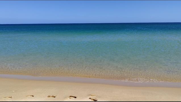 Snapshot of the sea at Meia Praia beach under strong North winds. The sea is flat, no waves and no surf, transparent over the wet sand and with several layers of turquoise blues towards the horizon where it becomes very dark blue. Above, clear baby blue skies. Along the shoreline several footsteps are seen in the wet sand.