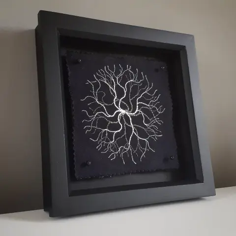 A black picture frame with a square piece of embroidery. White & grey threads depict a branching pattern emerging from the center.