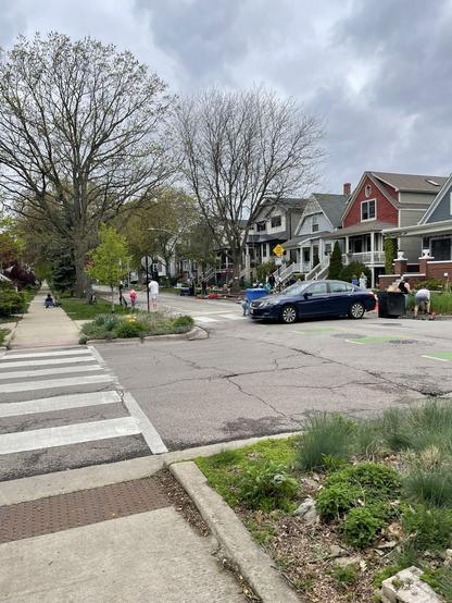 A personal car blocks a one way residential street. Children play in the street beyond. The trees are just beginning to show leaves, the sky is overcast.