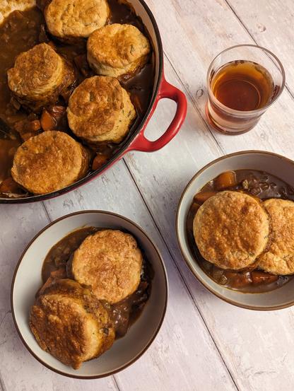 A photo of some food and a drink on a white wood-effect background. At the front are two bowls containing a dark brown stew with vegetables and beef, each with two golden American-style biscuits on top. To the top left is a shallow red cast-iron dish the cobbler was cooked in - it contains more stew and biscuits on top. To the top right is a low-ball glass with a dark brown drink in it.