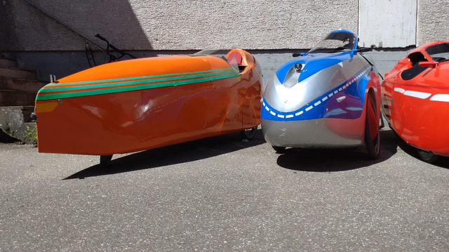 Thre velomobiles, an orange one with green belt lines and the for velomobiles very seldom delta layout, one with German police livery (said to have belonged to a policeman) and (just half in the picture, a red one with a white belt.