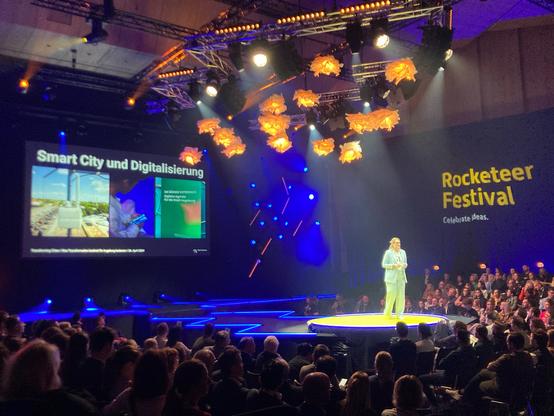 Oberbürgermeisterin Eva Weber in ihrer Keynote. Sie trägt einen hellblauen Anzug und steht in der Augsburger Kongresshalle umgeben von vielen Menschen . Hinter ihr ist eine Präsentation zu sehen. Sie spricht über Innovation und auch Smart City Augsburg.