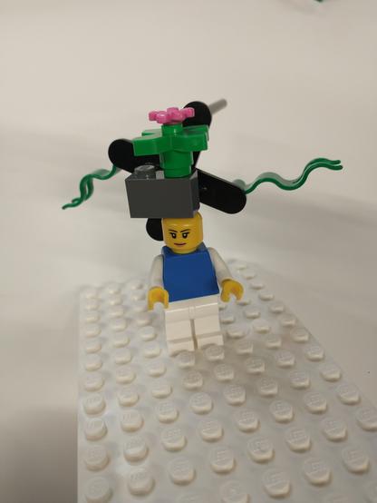 Eine Legofigur mit einem grauen 2er Block auf dem Kopf auf dem eine Pflanze angedeutet ist. Daran hängt ein Ventilator aus Lego.

All das auf einem weißen flachen Grundboard