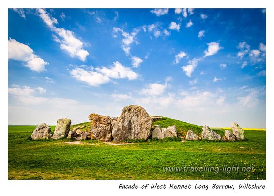 Photo of a row of upright stones arranged with the tallest in the middle to form a frontage to the end of a long mound, covered and surrounded by short grass.