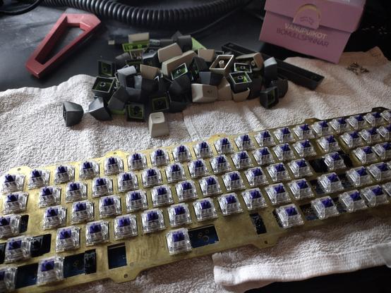 Photo of the PCB, plate, switches, and keycaps of a disassembled UK78 custom DIY mechanical keyboard. The plate-PCB-switch assembly has been detached from its aluminum case and is laying on a towel waiting for cleaning. The switches on the brass plate are purple stemmed clear-case Zealios switches.