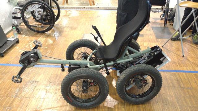 A pale green cargo recumbent quadcycle with a bottom/front bracket engine and fat, knobby tires. The rear third is folded below the seat.