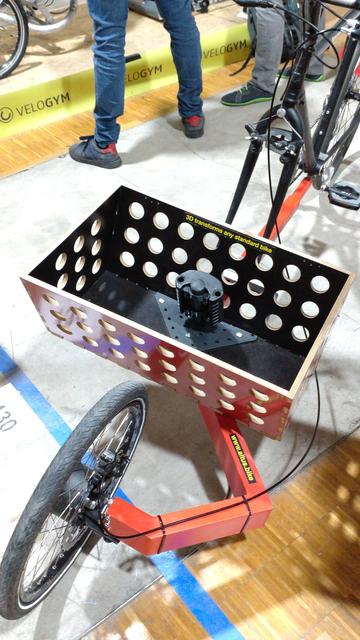 Front view of an old road cycle with a red cargobike front extension and a wooden (or MDF) cargo bay/basket with three rows of round holes and a standardized child seat mount point.