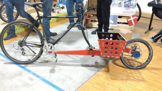 Profile view of an old black road cycle with a red cargobike front extension and a wooden (or MDF) cargo bay/basket with three rows of round holes.
