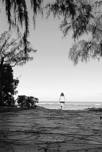 Je marche au loin vers la plage depuis un chemin d'accès en bois. Lagon au loin, végétation autour, ciel dégagé.
Photo en noir et blanc.