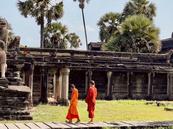 A couple young Buddhist monks, wearing orange robes, walk on a wooden walkway. Behind them the elevated stone walkway to the entrance of Angkor Wat runs parallel to them.