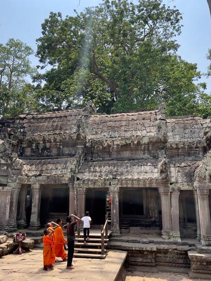 A group of 5 young boys, 3 of which are wearing the orange robes of novice monks, are entering the ruins at Ta Prohm. Three of the boys have stopped and are shielding their eyes from the sun as they look high above the ruins to look at an immense ￼tree, off photo, that has grown out of the ruins.